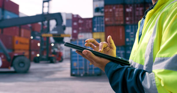 Worker using a tablet to manage shipping containers at a busy freight yard.