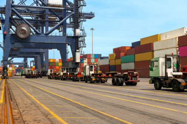 Container trucks lined up at a port terminal with stacked shipping containers and cranes.