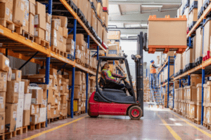 Busy pick and pack warehouse with workers using a forklift to move boxes along tall shelving aisles.