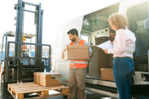 Workers unloading boxes from a delivery van while a forklift and pallet stand by during restocking. 
